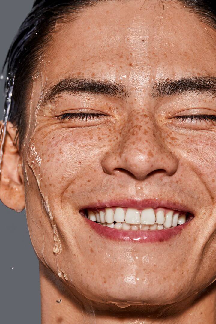 Close-up of a smiling face with hydrated skin and water droplets, representing IMEAN’s peptide-powered hydration effect.