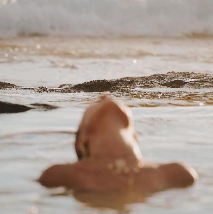 A person floating headfirst in shallow water, viewed from behind, with a blurred natural background of ripples and sunlight.