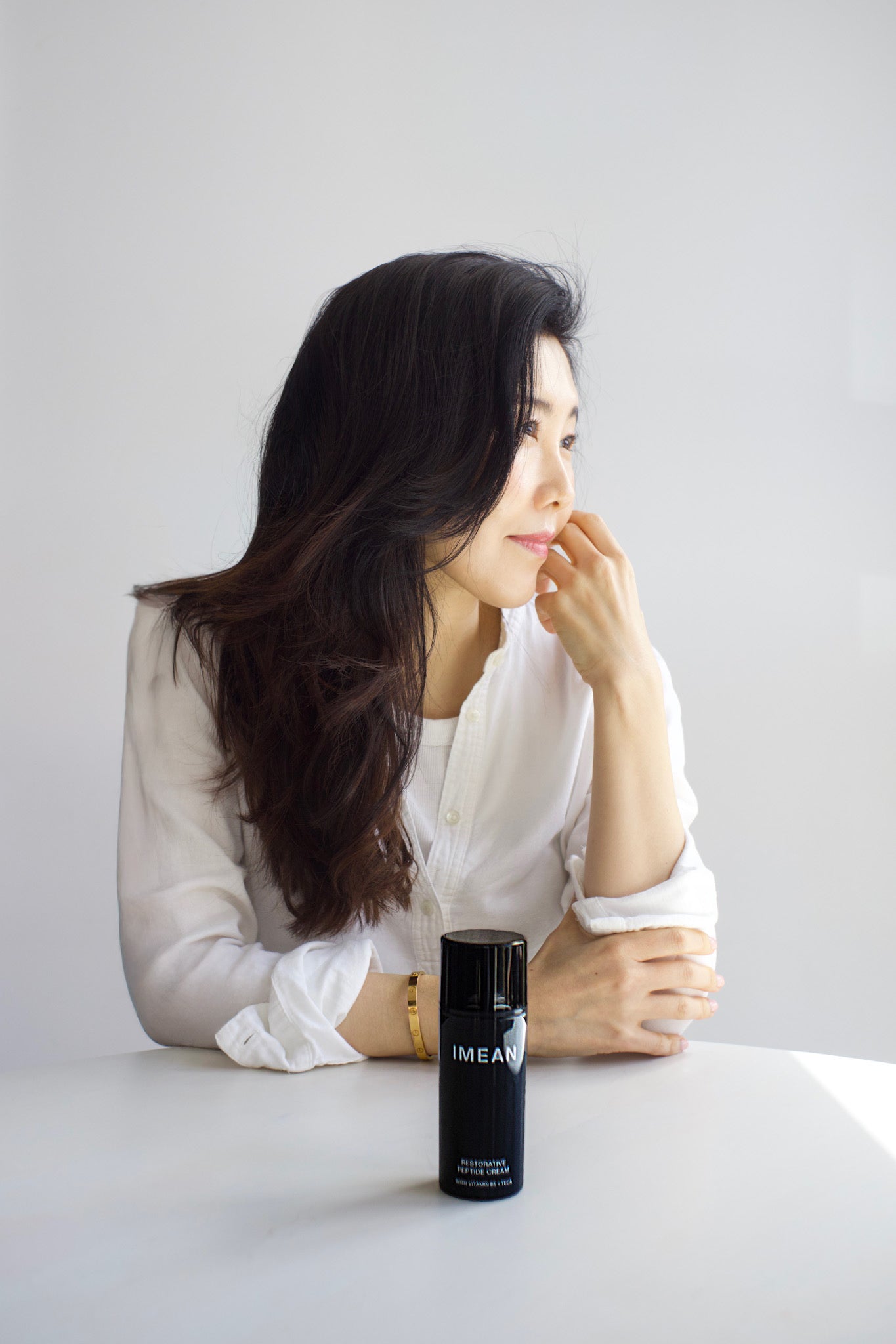 IMEAN founder Sarah Choi in a white shirt, resting her chin on her hand beside a bottle of IMEAN Restorative Peptide Cream
