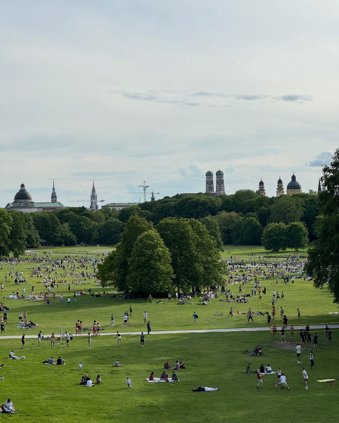 People enjoying a sunny afternoon on the lawn at Englischer Garten in Munich, with the city's iconic skyline in the background — a visual in Sarah’s Edit to Munich, Germany from IMEAN Beauty travel guide