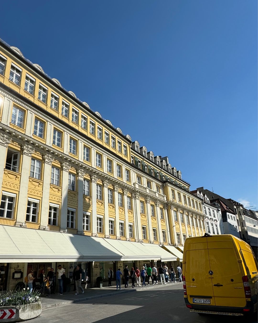 Historic yellow building in Munich’s old town under a clear blue sky, with a yellow van parked on the street and people gathered outside restaurants — part of IMEAN’s Munich Travel Guide.