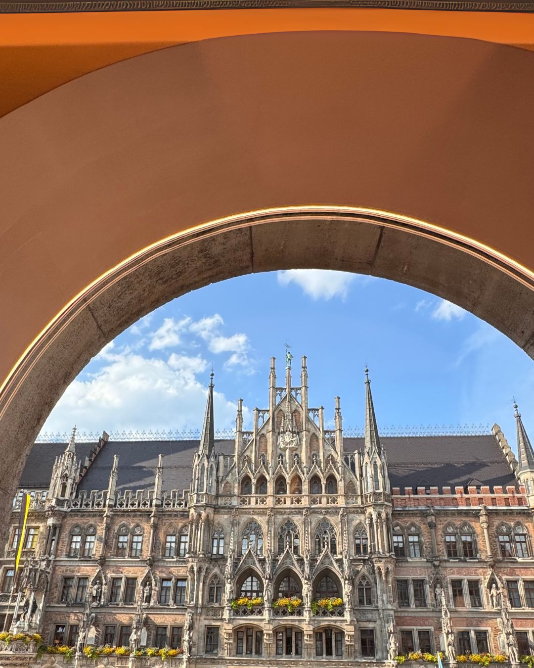 Neo-Gothic New Town Hall (Neues Rathaus) in Marienplatz, Munich, viewed through an orange-toned archway under a blue sky — a visual captured in Sarah’s Edit to Munich, Germany from IMEAN Beauty’s travel guide.