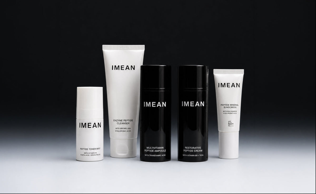 Five IMEAN skincare bottles and tubes arranged on a reflective surface against a dark studio background. The set includes white and black containers with minimalist typography.